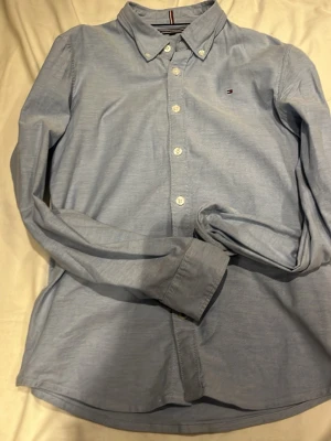 Ljusblå skjorta från Tommy Hilfiger - Snygg ljusblå skjorta från Tommy Hilfiger med klassisk button-down krage och diskret logga på bröstet. Skjortan har vita knappar och lång ärm, perfekt för en clean och stilren look. Materialet känns mjukt och är troligen bomull.