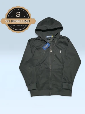 Svart hoodie från Polo Ralph Lauren - Snygg svart hoodie från Polo Ralph Lauren med dragkedja framtill och klassisk huva med dragsko. Ikoniska vita loggan broderad på bröstet. Tillverkad i mjukt bomullsmaterial och har två fickor framtill. Perfekt för en avslappnad och stilren look.