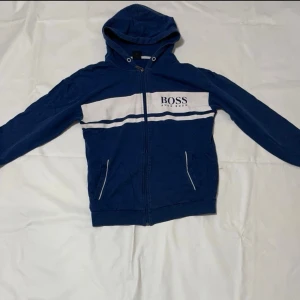 Blå hoodie från BOSS med dragkedja - Snygg blå hoodie från BOSS med vit panel och logga på bröstet. Hoodien har dragkedja framtill, två fickor och vit rand över bröstet. Klassisk huva och ribbade muddar. Perfekt för en avslappnad och trendig look.