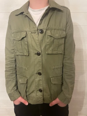 Olivgrön utilityjacka med fickor - Snygg olivgrön utilityjacka med fyra stora fickor framtill och knäppning med svarta knappar. A40, b40, c43, d60
