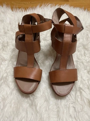 Marc oPolo  - Stilrena bruna pumps med breda remmar och öppen tå. Skorna har en stadig klack och justerbart spänne runt ankeln. Tillverkade i skinnliknande material och har en modern design som ger en snygg siluett.