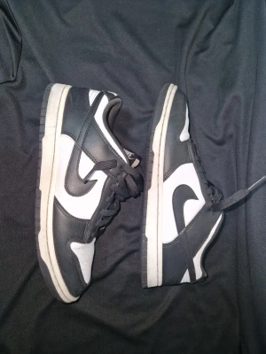 Nike Dunk Low svart/vit sneakers - Säljer ett par klassiska Nike Dunk Low sneakers i svart och vitt. Skorna har en låg siluett, rund tå, snörning och den ikoniska swoosh-loggan på sidan. Tillverkade i skinn med vit bas och svarta överlägg. Perfekta för dig som gillar streetwear och tidlös stil.