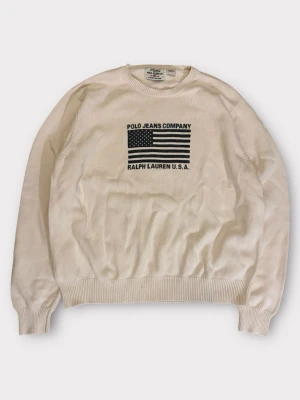 Vit stickad tröja Polo Ralph Lauren Jeans Company - Vit stickad tröja från Polo Jeans Company Ralph Lauren med svart amerikansk flagga och text på bröstet. Tröjan har rund halsringning, ribbade muddar och är tillverkad i mjukt material. Perfekt för en clean och klassisk look. Märkt XL Passar L