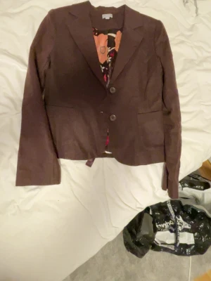 Brun manchesterkavaj med knytband - Snygg brun kavaj i manchester med två knappar framtill och dekorativa knytband bak. Klassisk krage och långa ärmar med knappdetaljer vid ärmslut. Perfekt för dig som vill ha en unik twist på din outfit.