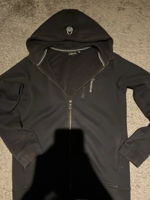 Svart hoodie från Sail Racing - Snygg svart hoodie från Sail Racing med dragkedja framtill och vit logga på bröstet samt huvan. Hoodien har långa ärmar, ribbade muddar och två fickor. Tillverkad i mjukt och bekvämt material, perfekt för en avslappnad stil.