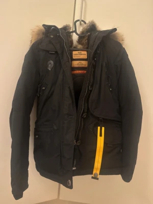 Svart Parajumpers parkas med päls - Säljer en svart Parajumpers parkas X small med stor huva och fluffig pälsdetalj. Jackan har flera fickor, dragkedja, knappar och den ikoniska gula remmen. Perfekt för kalla vinterdagar och riktigt snygg streetstil.