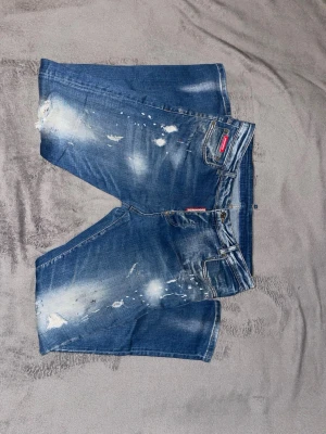 Blå Dsquared2 jeans med slitningar - Säljer ett par blå jeans från Dsquared2 med coola slitningar och färgstänk på framsidan. Shortsen har klassisk femficksdesign, tydliga slitna detaljer och en röd Dsquared2-logga vid fickan. Perfekta för en avslappnad streetwear-look.