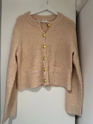 Beige kofta från & Other Stories - Mysig beige kofta från & Other Stories med guldknappar framtill och två fickor. Tröjan är stickad i ett fluffigt material och har lång ärm samt ribbade detaljer vid krage och nederkant. Perfekt för lager på lager under kyliga dagar.