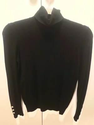 Svart polotröja med guldknappar - Säljer en svart polotröja med lång ärm och snygga guldknappar vid ärmsluten. Tröjan har en hög krage och är i mjukt, stickat material. Perfekt för dig som vill ha en clean och stilren look med en liten twist. Tyvärr är tröjan lite noppig