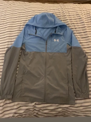 Blågrå vindjacka från Under Armour - Under armour jacka i blå. Bara en liten fläck där bak som man inte ser alls om man tittar på ser bara om du tittar någrant. Skriv ifall du vill ha fler bilder och kan vid snabba affärer så kan priset variera och ett litet hål vid armen