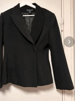 Svart kavaj från H&M med klassisk krage - Stilren svart kavaj från H&M med klassisk krage och figurnära passform. Kavajen har långa ärmar, enkel knäppning framtill och är helfärgad utan mönster. Perfekt för en clean och snygg look.