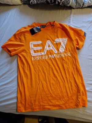 Orange EA7 Emporio Armani t-shirt - Snygg orange t-shirt från EA7 Emporio Armani med stort vitt tryck på bröstet. Klassisk rund hals och korta ärmar. Perfekt för dig som vill sticka ut med en färgstark och trendig look. Tillverkad i mjuk bomull för skön känsla. Tags sitter kvar, aldrig använd.