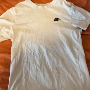 Vit t-shirt från Nike - Klassisk vit t-shirt från Nike med liten svart logga på bröstet. Enkel och stilren design med rund hals och korta ärmar. Perfekt till jeans eller shorts för en clean look.