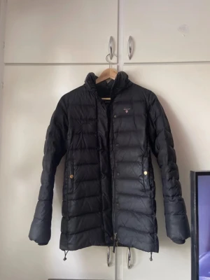 Svart dunjacka från Gant XS - Snygg svart dunjacka från Gant i storlek XS. Jackan har quiltad design, dragkedja och tryckknappar framtill samt två fickor med guldfärgade knappar. Har varit ett hål fram men har sytt den men inget man tänker på:) skulle behöva en kemtvätt 