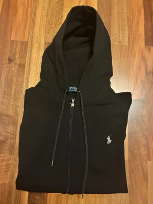 Svart hoodie från Polo Ralph Lauren - Snygg svart hoodie från Polo Ralph Lauren med dragkedja framtill och klassisk huva med snören. Ikoniska vita loggan broderad på bröstet. Tillverkad i mjukt bomullsmaterial som känns skönt mot huden. Perfekt för en avslappnad och stilren look.