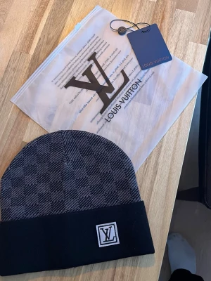 svart grå mönstrad mössa - Snygg svartgrå mössa från med klassiskt rutmönster och vikt kant. logga framtill på kanten ger en exklusiv touch. Tillverkad i mjukt stickat material som sitter skönt på huvudet. Perfekt accessoar för dig som gillar lyxiga detaljer.