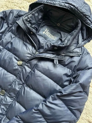 Mörkblå dunjacka från Herno - Säljer en mörkblå dunjacka från Herno med huva och både dragkedja och knappar framtill. Jackan är quiltad och har en klassisk, stilren look som passar perfekt när det är kallt ute. Fodrad och riktigt skön att bära under höst och vinter. Modell  185 cm!