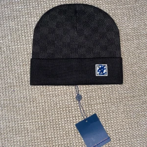 Svart mössa - Snygg svart mössa med diskret rutmönster och vikt kant. På framsidan sitter en patch med blått monogram. Perfekt accessoar för att lyfta din look under kyliga dagar.