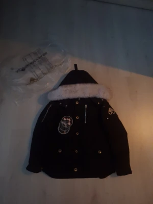 Svart dunjacka Moose Knuckles - Säljer en svart dunjacka från Moose Knuckles med stor vit fuskpäls runt huvan och coola patches. Jackan har dragkedjor, silvriga detaljer och flera fickor. Perfekt för kalla dagar och riktigt snygg streetstil.