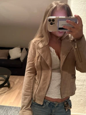 Beige mockajacka med dragkedjor - Säljer en snygg beige mockajacka med coola dragkedjor och klassisk krage. Jackan har en figurnära passform och är perfekt för dig som vill ha en stilren look. Passar till jeans och ger en trendig vibe.