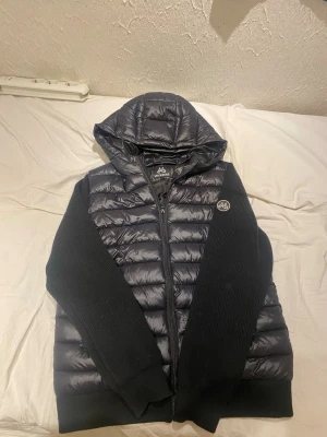 Svart pufferjacka från Mountain Works - Snygg svart pufferjacka från Maggiore med quiltad kropp och stickade ärmar. Jackan har huva, dragkedja framtill och en rund logotyp på bröstet. Perfekt för kyliga dagar när du vill ha både stil och komfort.