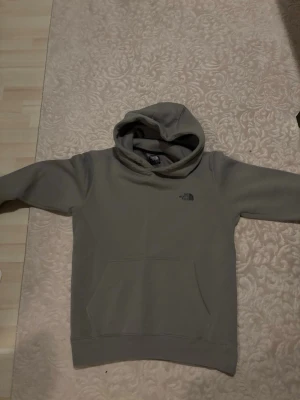 Mörk grå hoodie från The North Face - Snygg beige hoodie från The North Face med klassisk logga på bröstet. Hoodien har stor känguruficka framtill, ribbade muddar och en rejäl huva. Perfekt för chill dagar och streetwear-stil. Mjuk och skön bomullsblandning.