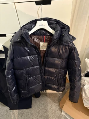 Mörkblå dunjacka från Moncler - Snygg mörkblå dunjacka från Moncler med quiltad design och avtagbar huva. Jackan har dragkedja framtill, två sidofickor med dragkedja och en ikonisk Moncler-logga på ärmen. Insidan har ett unikt serietryck och innerficka med dragkedja. Storlek 3