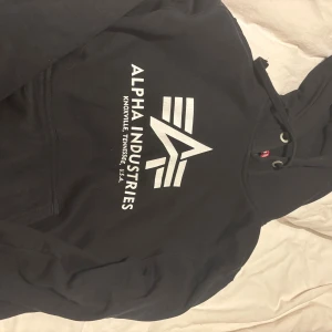 Svart hoodie från Alpha Industries - Snygg svart hoodie från Alpha Industries med stor vit logga och text på bröstet. Klassisk huva med dragsko och mjukt material som känns skönt mot huden. Perfekt för en avslappnad streetwear-look.