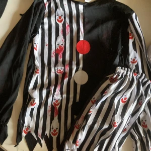 Randig clownkostym med tryck - Säljer en clownkostym i svart och vitt med randigt mönster och tryck av röda clownansikten. Kostymen har långa ärmar, elastisk midja på byxorna och två stora cirklar framtill i rött och vitt. Materialet är 100% polyester, perfekt för halloween eller temafest.