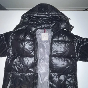 Svart pufferjacka från Moncler - Säljer en svart, glansig pufferjacka från Moncler med huva och dragkedja. Jackan har Moncler-logga på ärmen, stora fickor och quiltad design. Fylld med vitt gåsdun för extra värme. Perfekt för kalla dagar och riktigt snygg streetstil.