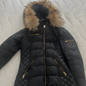 Svart Rockandblue pufferjacka med djurpäls  - Svart pufferjacka från Rockandblue med snygga guldfärgade dragkedjor och quiltad design. Jackan har en stor huva med fuskpäls och flera fickor med dragkedja. Märkespatch på ärmen och figurnära passform. Perfekt för kalla dagar.litet hål på armen syns inte