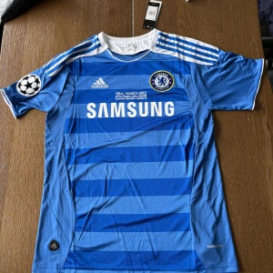 Chelsea CL 2012 Drogba tröja Adidas - Chelsea FC Champions League 2012 finaltröja med Drogba 11 på ryggen. Blå med vita detaljer, Adidas-logga, klubbmärke och Champions League-märke på ärmen. Tillverkad i lätt och ventilerande polyester (Climacool). Klassisk randig design och Samsung som sponsor.