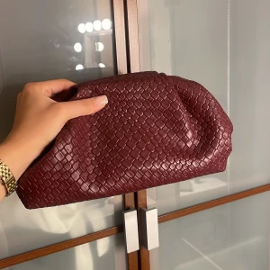 Vinröd kuvertväska i flätat skinn - Säljer en vinröd kuvertväska i flätat skinn med mjuk, puffig form. Väskan har en elegant och trendig look som passar perfekt till din outfit. Den är rymlig nog för det viktigaste och har en lyxig känsla tack vare det flätade mönstret.