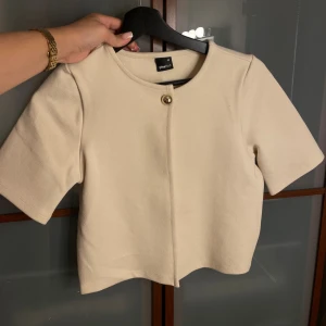 Beige kort tröja från Gina Tricot - Kort beige tröja från Gina Tricot med rund halsringning och en snygg guldig knapp framtill. Tröjan har korta ärmar och en stilren, minimalistisk look som är lätt att matcha. Perfekt för dig som gillar clean och modern stil. Den har endast en knapp vilket gör den öppen längst ner. 