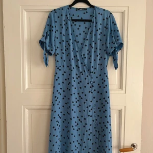 Blommig blå omlottklänning från Gina tricot - Supersöt blå långklänning från Gina tricot med svarta blommor och omlottmodell. Klänningen har korta ärmar med knyt och en v-ringad hals. Perfekt för sommarens alla tillfällen och ger en chill men ändå snygg vibe.