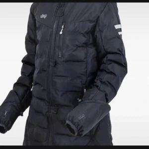  lång dunjacka från Uhip - Lång svart dunjacka från Uhip med dragkedja framtill, hög krage och reflekterande detaljer på ärmar och rygg. Jackan har flera praktiska fickor och är perfekt för kalla vinterdagar. Snygg och funktionell design som håller dig varm. Ett litet hål vid armhålan som inte syns på utsidan ❤️