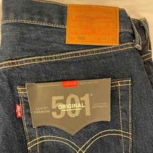Levi's 501 Original jeans blå W34 L30 - Klassiska Levi's 501 Original jeans i mörkblå denim med raka ben och signaturknappgylf. Snygga kontrastsömmar och ikonisk läderpatch bak. Premiumkvalitet och tidlös stil, perfekta till sneakers eller boots. Levi’s Premium 501 Original – klassiska raka jeans i mörk denim, oanvända med tag kvar.