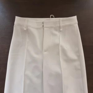 Vita kostymbyxor med pressveck - Snygga vita kostymbyxor med raka ben och pressveck framtill. Byxorna har hällor för bälte och stängs med två hakar och dragkedja. Materialet är slätt och känns dressat, perfekt för en clean och stilren look. 