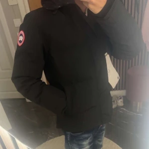 Svart dunjacka från Canada Goose - Säljer en svart dunjacka från Canada Goose med klassisk patch på ärmen och huva. Jackan har dragkedja och raka fickor framtill. Perfekt för kalla dagar och riktigt snygg till jeans eller cargo. Stilren och enkel design som funkar till det mesta.