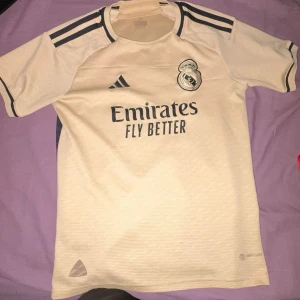Real Madrid Adidas fotbollströja beige - Snygg Real Madrid fotbollströja från Adidas i beige med mörkblå detaljer och klubbmärke på bröstet. Tröjan har korta ärmar, rund hals och är tillverkad i lätt, ventilerande AEROREADY-material. Perfekt för dig som älskar fotboll och vill visa ditt stöd för Real.