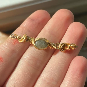 Guldarmband med grå sten (labradorit) - Unikt armband med guldfärgad kedja och snirkliga detaljer runt en oval grå sten i mitten. Låset är justerbart med en kedja för perfekt passform. Stenens naturliga mönster ger armbandet en cool och personlig vibe.