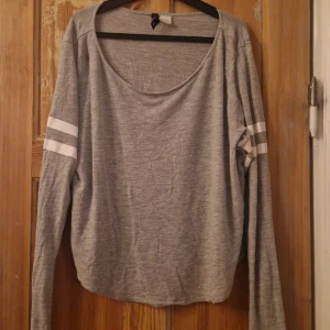 Grå långärmad tröja med vita ränder - Grå långärmad tröja med två vita ränder på varje ärm. Tröjan har en avslappnad passform och rundad halsringning. Perfekt för en chill och sportig look.