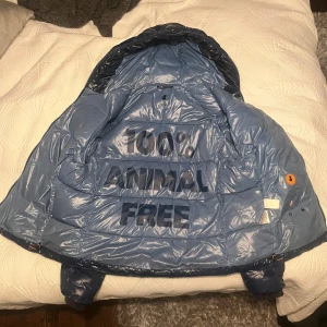 Blå pufferjacka från Save The Duck - Säljer en glansig blå pufferjacka från Save The Duck med huva och dragkedja. Jackan är perfekt för kalla dagar och är riktigt snygg. Har bara använt jackan en vinter och den har inga defekter eller hål. 