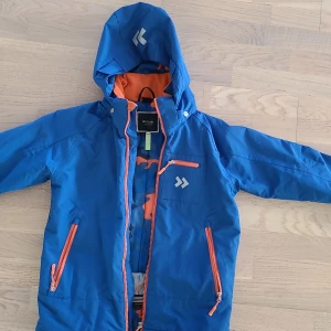 Blå vindjacka från 157 Funk, stl 150 - Blå vindjacka från 157 Funk med orange detaljer och dragkedjor. Jackan har huva, flera fickor med dragkedja och coolt camo-mönstrat foder i blått och orange. Perfekt för utomhusaktiviteter och har en sportig vibe.