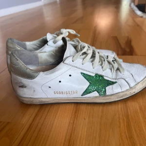 Golden Goose Superstar sneakers med grön stjärna - Golden Goose Superstar sneakers i vitt skinn med glittrig grön stjärna på sidan och silverfärgade detaljer vid hälen. Klassisk rund tå, snörning och platt sula. Snyggt slitna för en cool streetstil. Perfekt för dig som gillar unika och trendiga sneakers.
