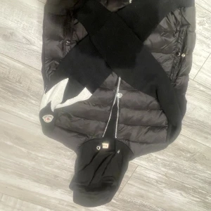 Svart Moncler dunjacka med huva - Snygg svart dunjacka från Moncler med quiltad design, huva och dragkedja framtill. Jackan har Moncler-logga på ärmen och praktiska fickor med knappar. Perfekt för kalla dagar och ger en clean, modern look.