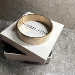 Bredt armband från Dyrberg/Kern i guld - Stilrent och brett armband från Dyrberg/Kern i guldig metall med en slät, blank yta och diskret logga på sidan. Perfekt accessoar för att lyfta din outfit med en lyxig touch. Kommer i originalask. Diameter: 6,5 cm Bredd: 2 cm