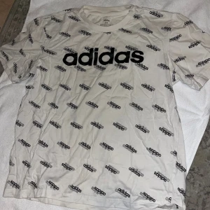 Tshirt - Vit t-shirt från Adidas med svart logotyptryck över hela plagget och stor Adidas-logga på bröstet. Klassisk rund hals och korta ärmar. Tillverkad i mjuk bomull, perfekt för en sportig och avslappnad stil. En liten fläck jag inte provat ta bort