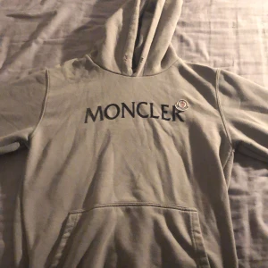 Beige Moncler hoodie med logga - Säljer en beige hoodie från Moncler Grenoble med stor svart logotyp på bröstet och klassisk Moncler-patch. Tröjan har huva, känguruficka och långa ärmar. Perfekt för dig som gillar exklusiva streetwear-vibbar.