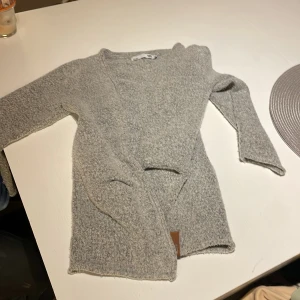 Grå stickad kofta från 157 - Mysig grå stickad kofta från 157 med öppen front och lång ärm. Koftan har en enkel och stilren design, perfekt att slänga på sig när det är kyligt. Materialet är mjukt och ger en avslappnad vibe.